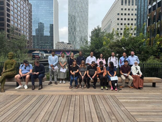 A group of people with visual impairments and their guides sitting on a bench in Canary Wharf, London.